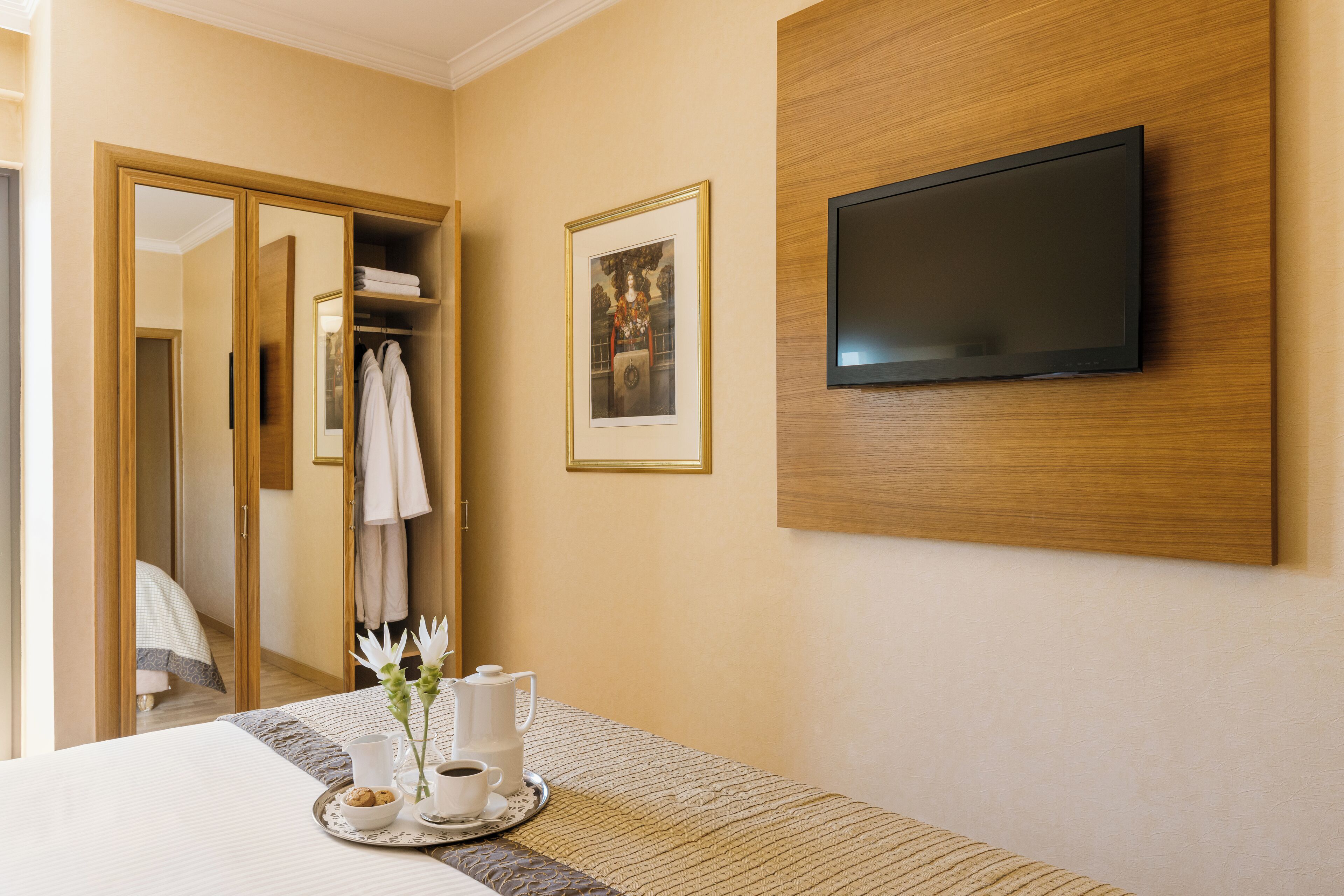 Habitación de hotel con una cama, un televisor montado en la pared, un cuadro enmarcado y una bandeja con cafetera y galletas.