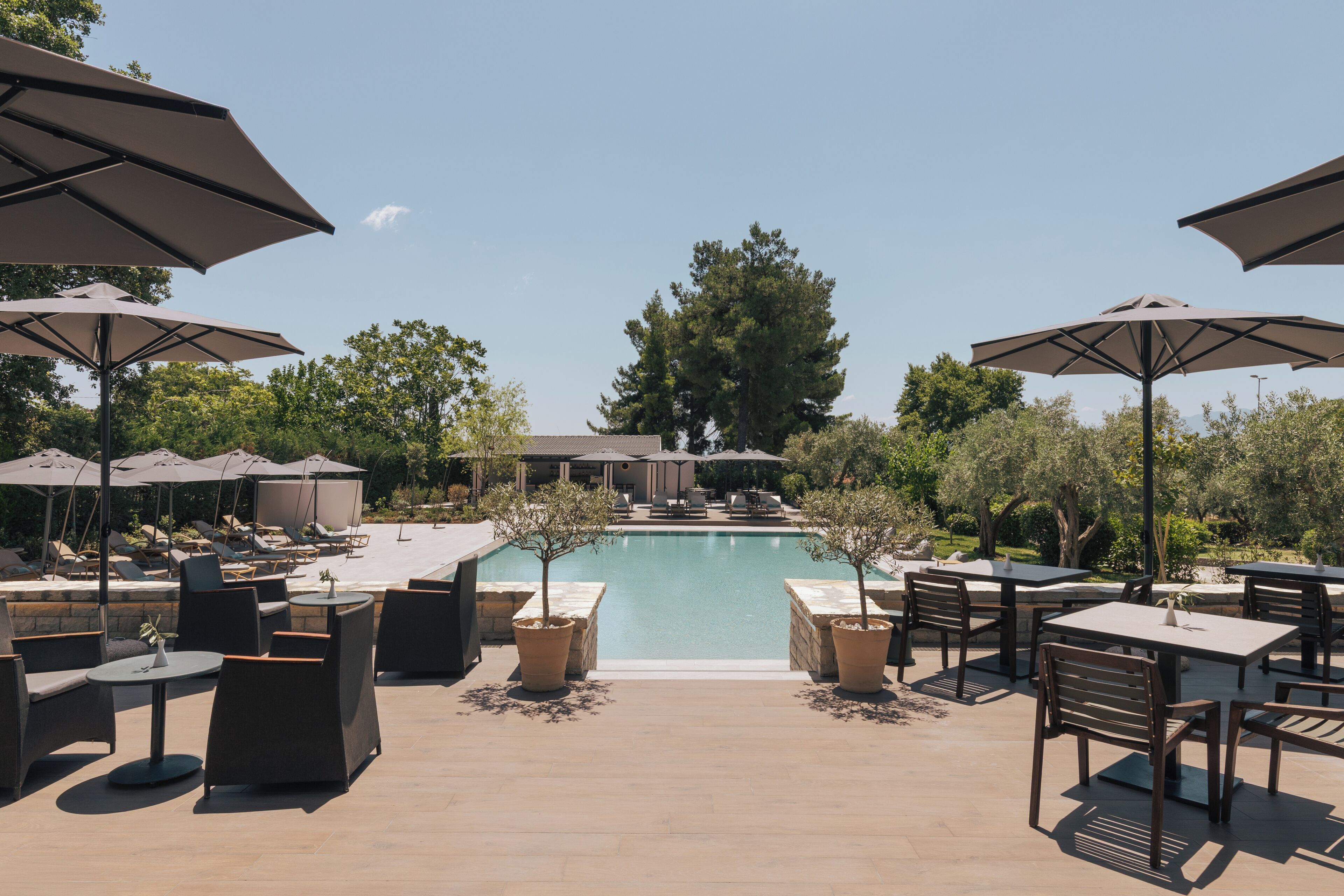 An outdoor pool area with lounge chairs and umbrellas, surrounded by trees and a building in the background.