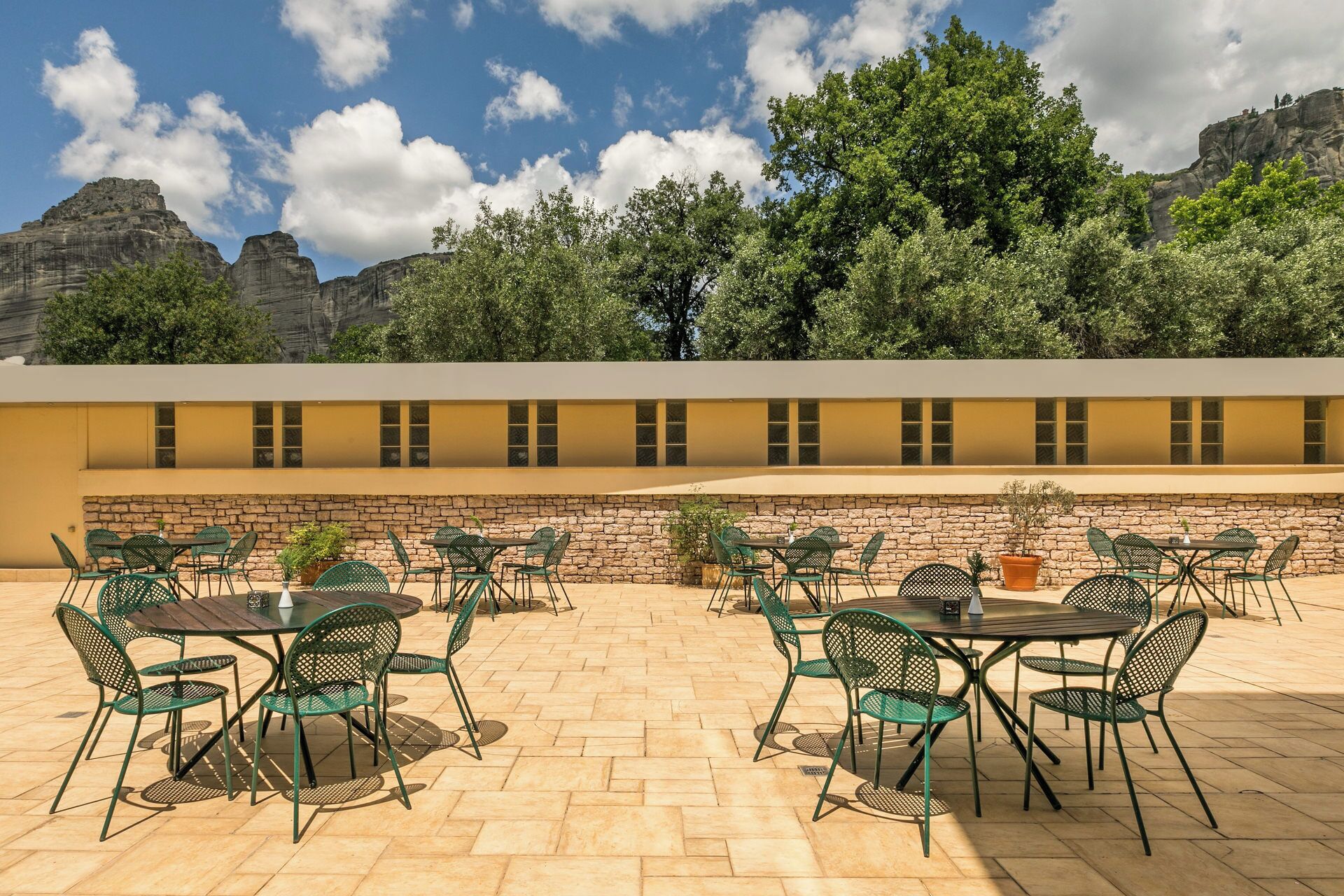 An outdoor patio with multiple tables and chairs, a stone wall, and a view of mountains and trees.
