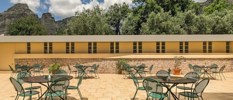 Une terrasse extérieure dotée de plusieurs tables et chaises, d’un mur en pierre et offrant une vue sur les montagnes et les arbres.