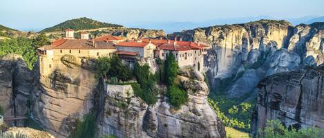 A historic building perched on a cliff with multiple levels and a red-tiled roof, surrounded by rugged cliffs and greenery.