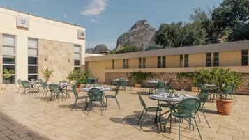 An outdoor patio with tables and chairs, a building with large windows, and a mountain in the background.