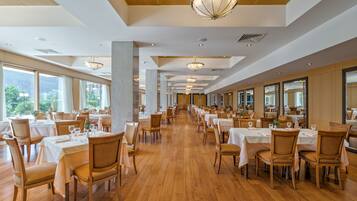 A spacious dining area with long tables and chairs, large windows, and a high ceiling with chandeliers.