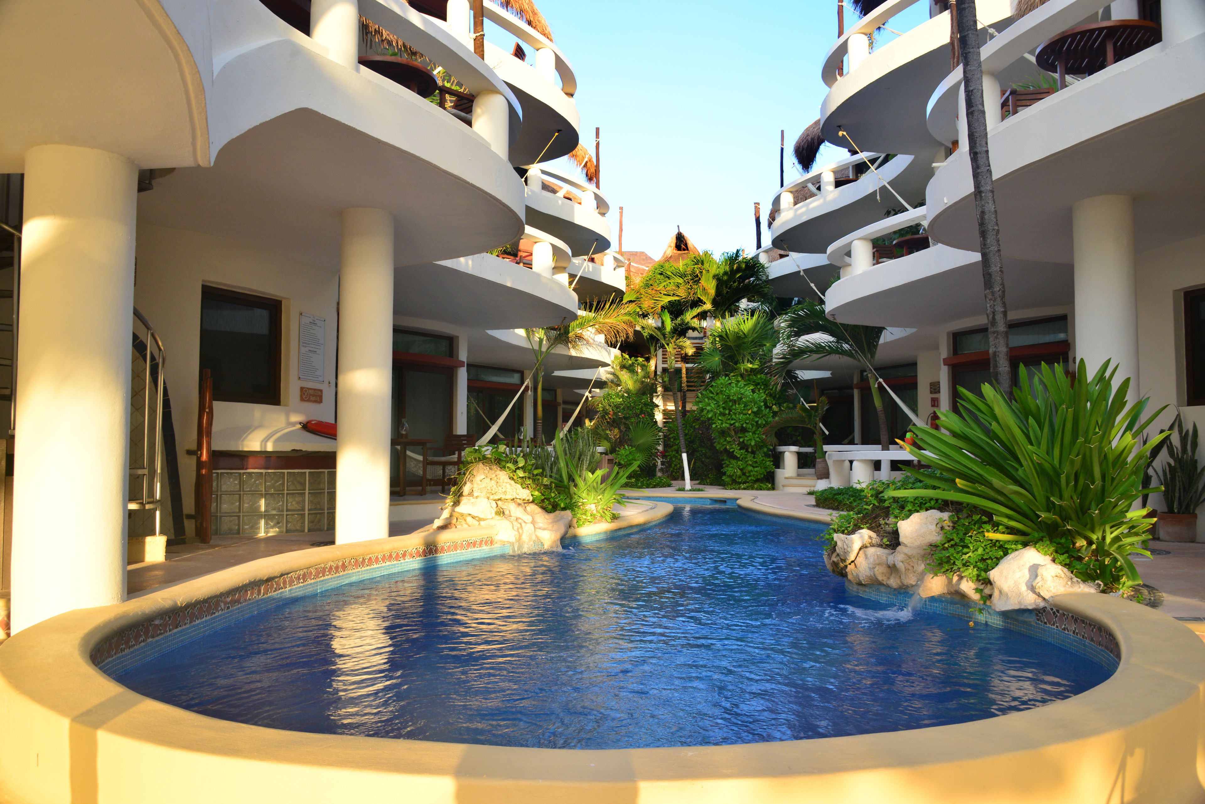 Une piscine dont les bords sont bordés de rochers, entourée de palmiers et d’un immeuble à plusieurs niveaux doté de balcons.