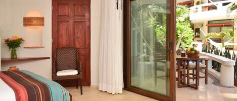 Una habitación con una cama, una silla, una puerta de madera, una puerta corrediza de vidrio que da a un balcón con plantas y una mesa.