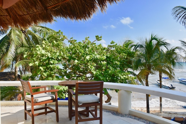 Una mesa y sillas de madera en un balcón con vistas a la playa y palmeras.