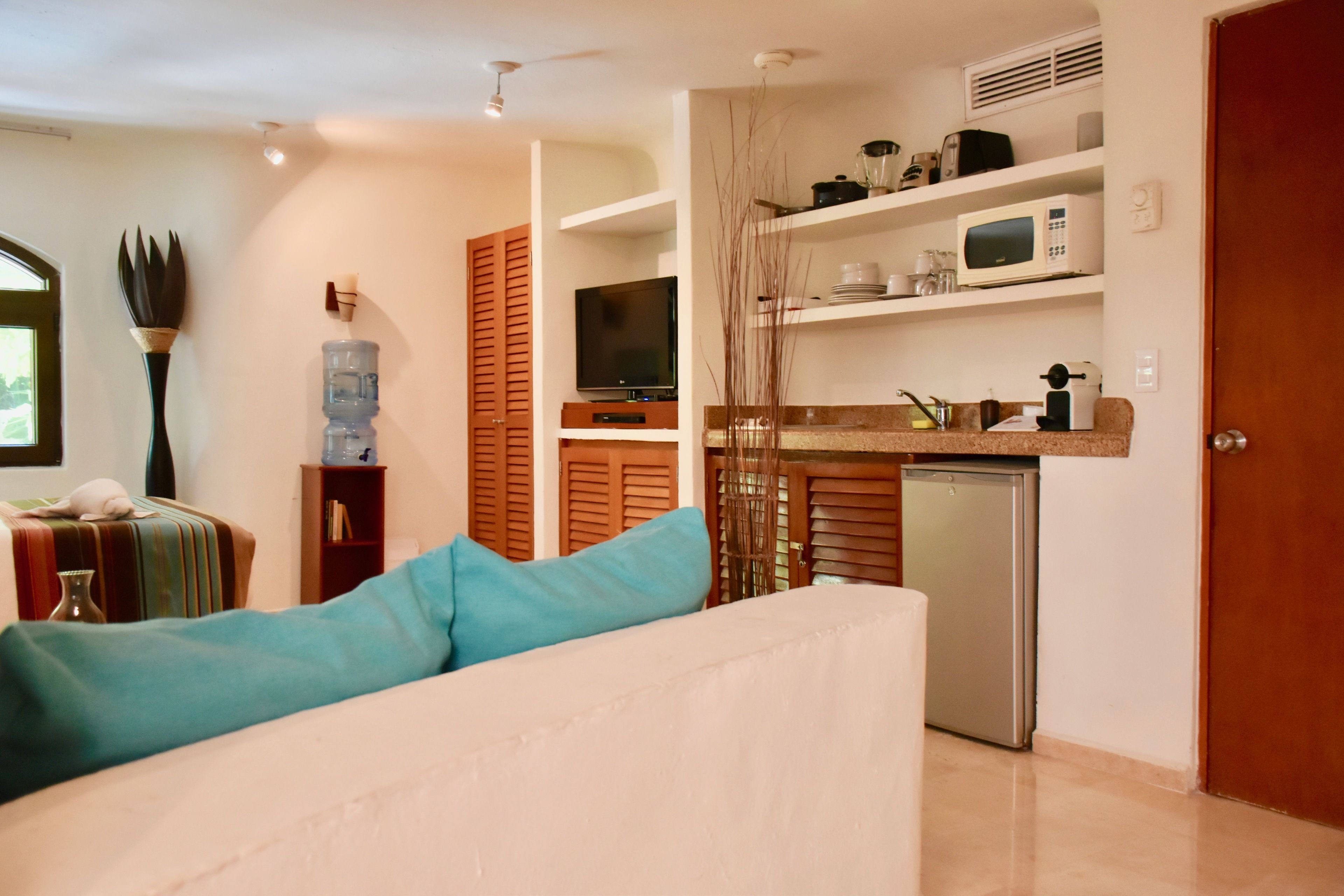 A modern living room with a sofa, a water dispenser, a television, and a kitchen area with a microwave and a refrigerator.