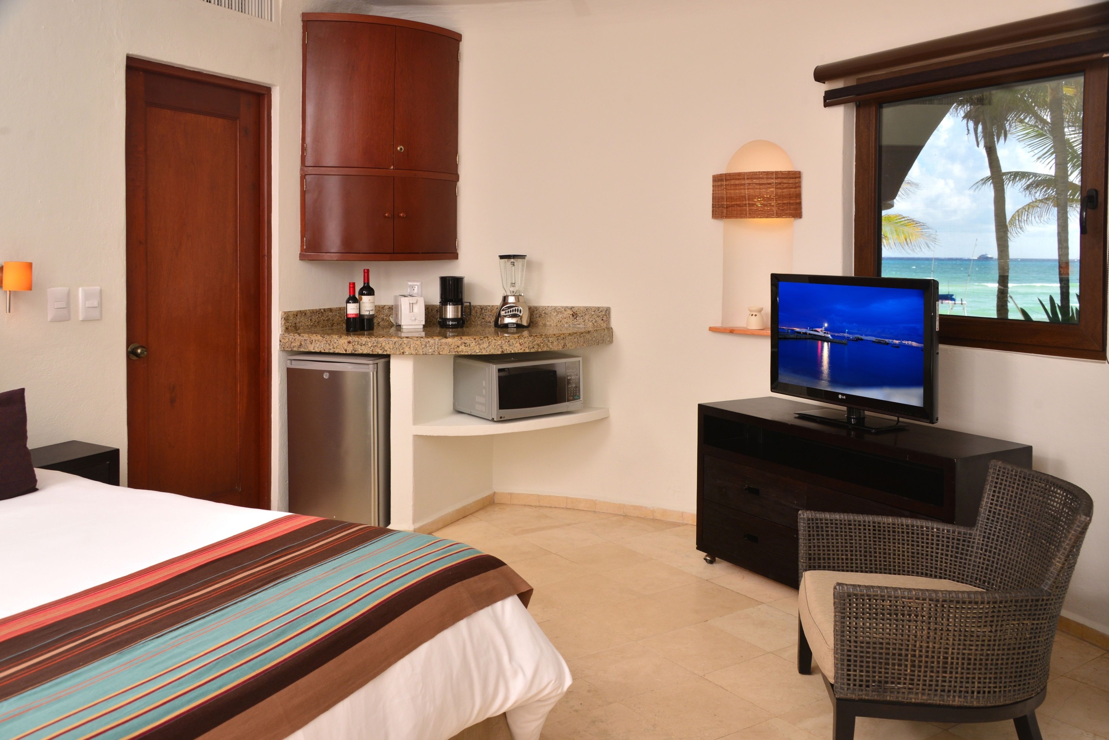 Ein Hotelzimmer mit Bett, Fernseher, Mikrowelle, Kühlschrank, einer kleinen Küchenzeile, einem Korbstuhl und Meerblick durch ein Fenster.
