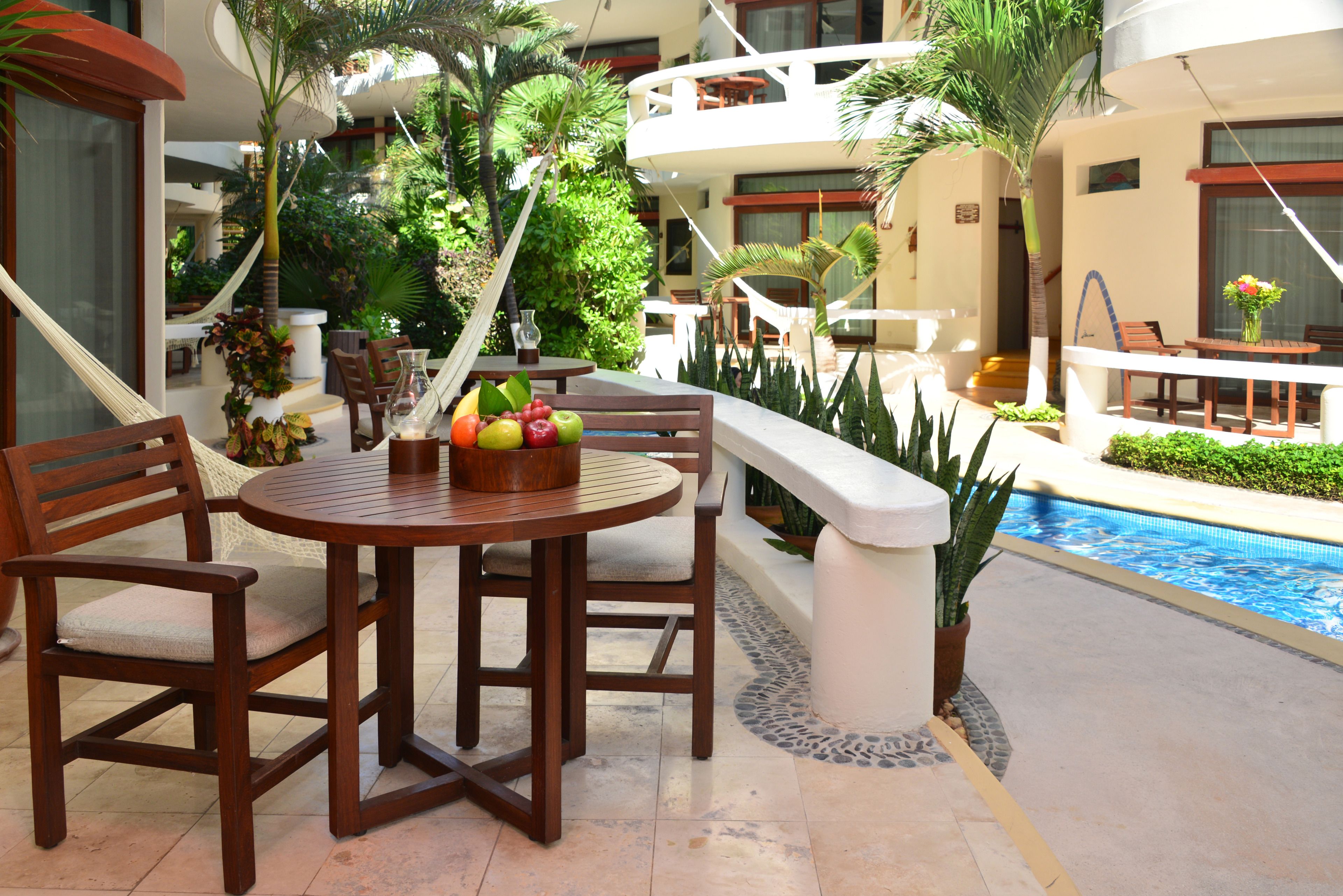Un espace aménagé au bord de la piscine, avec une table ronde en bois et des chaises, entouré d’une végétation luxuriante et donnant sur une piscine.