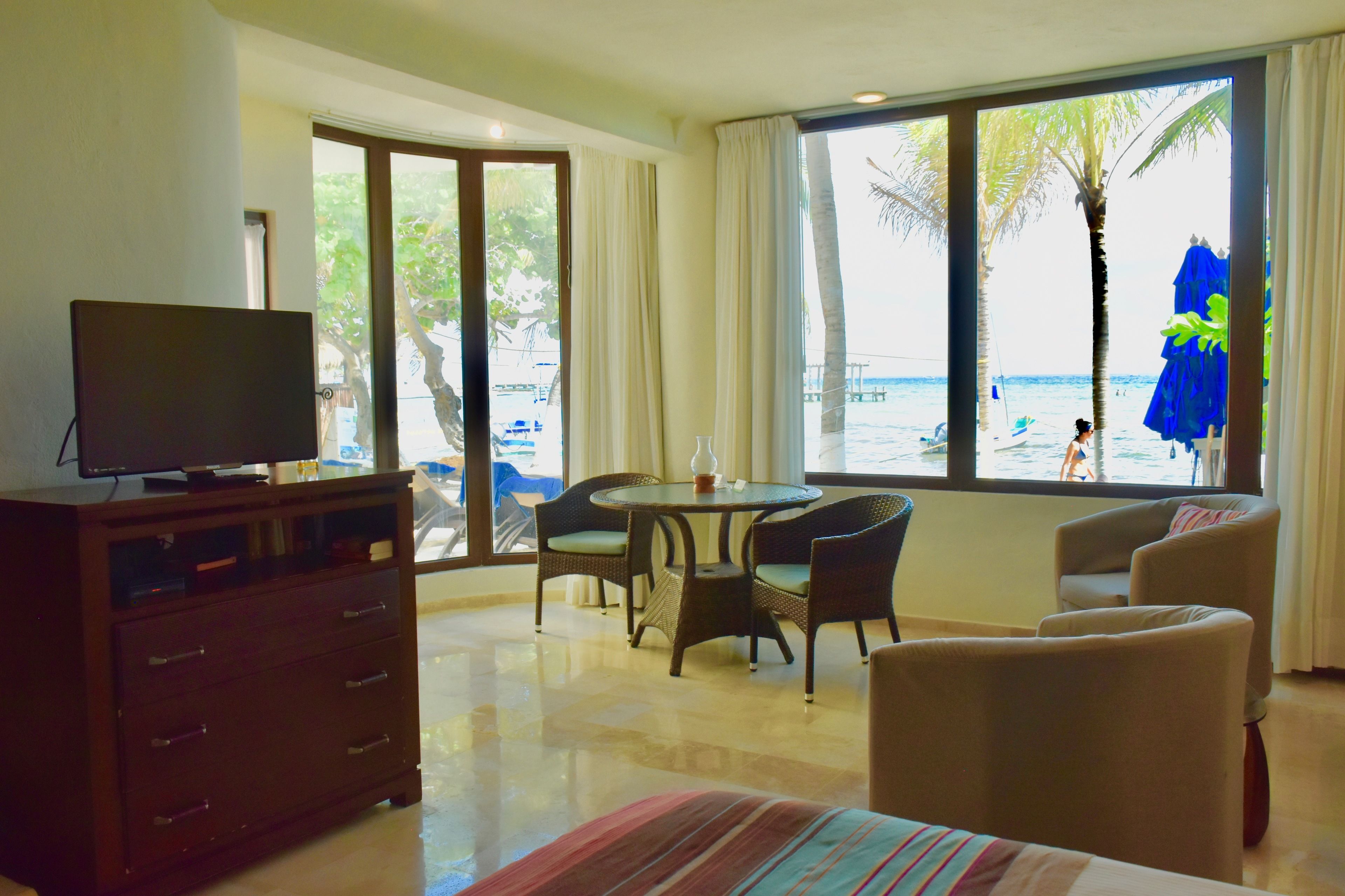 A hotel room with a television on a dresser, a dining table with chairs, a sofa, and a large window with a view of the beach and palm trees.