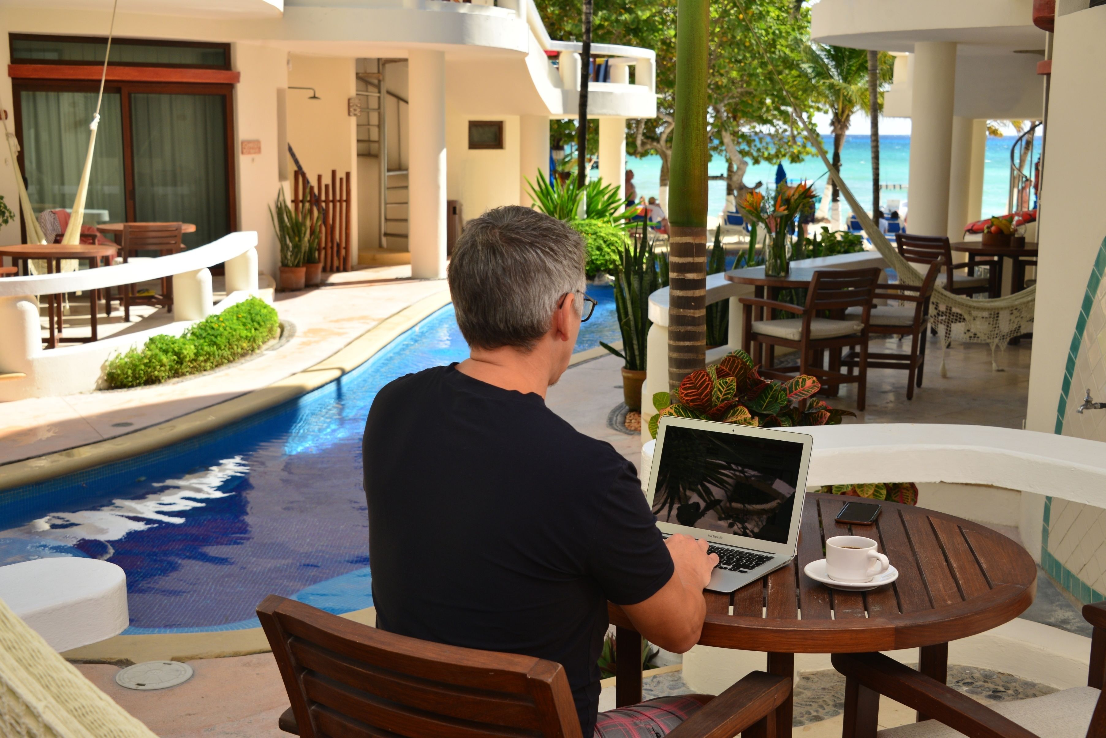 Un hombre usando una computadora portátil en una mesa de madera junto a una piscina, con vista al océano.