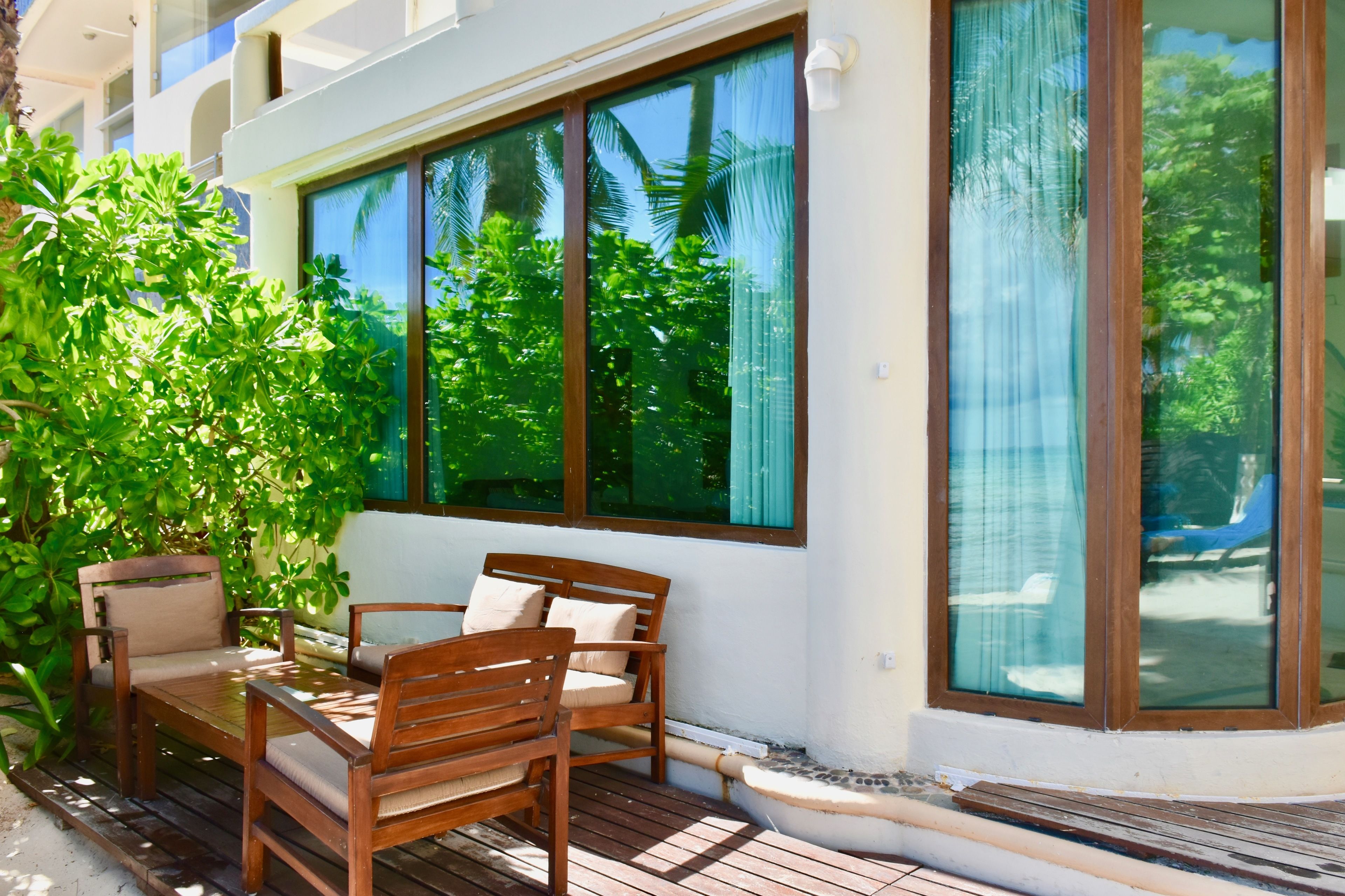 A wooden deck with a table and chairs, large windows, and lush greenery.