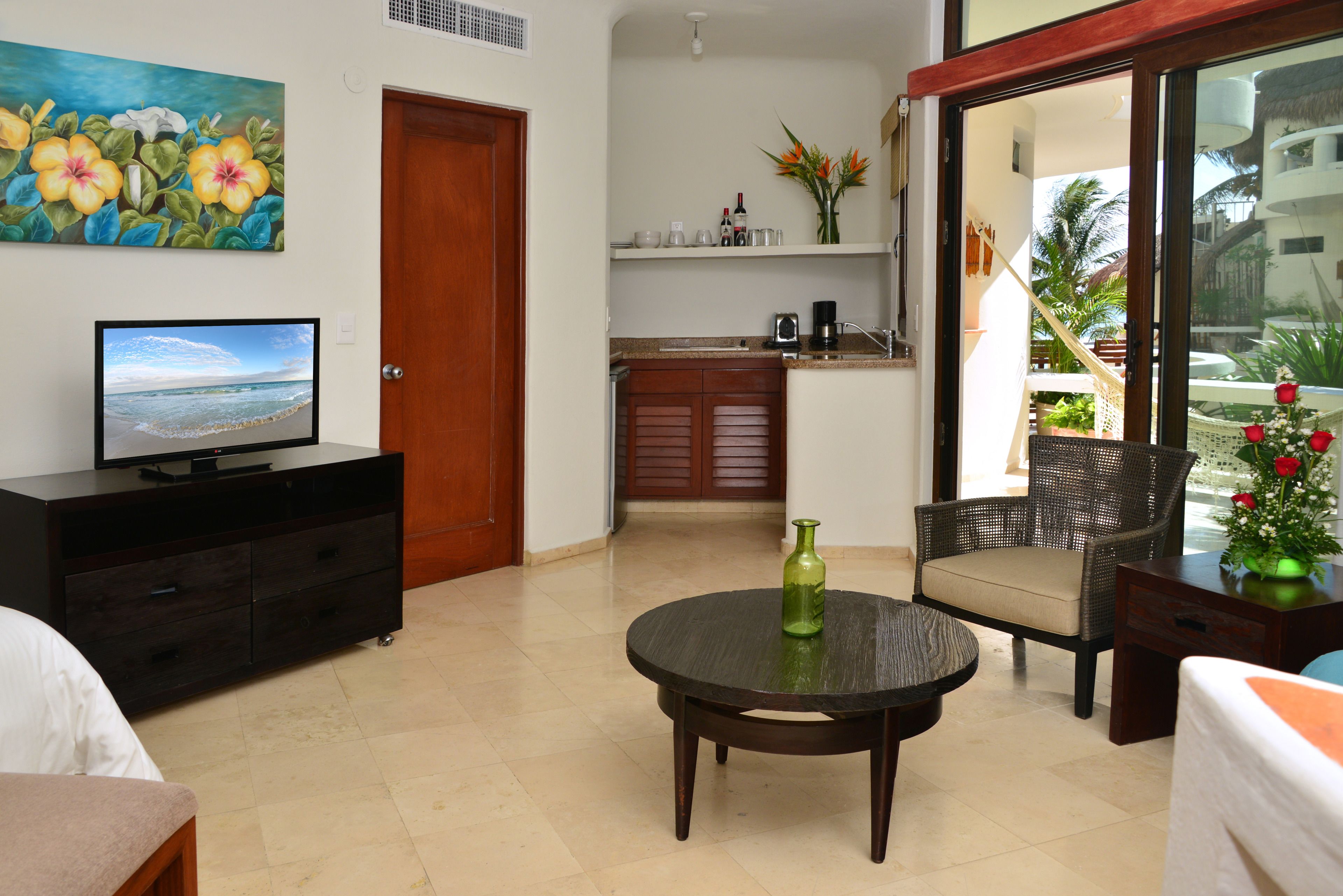 A hotel room with a television, a dining table, a chair, a small kitchen area, and a view of the outside balcony.