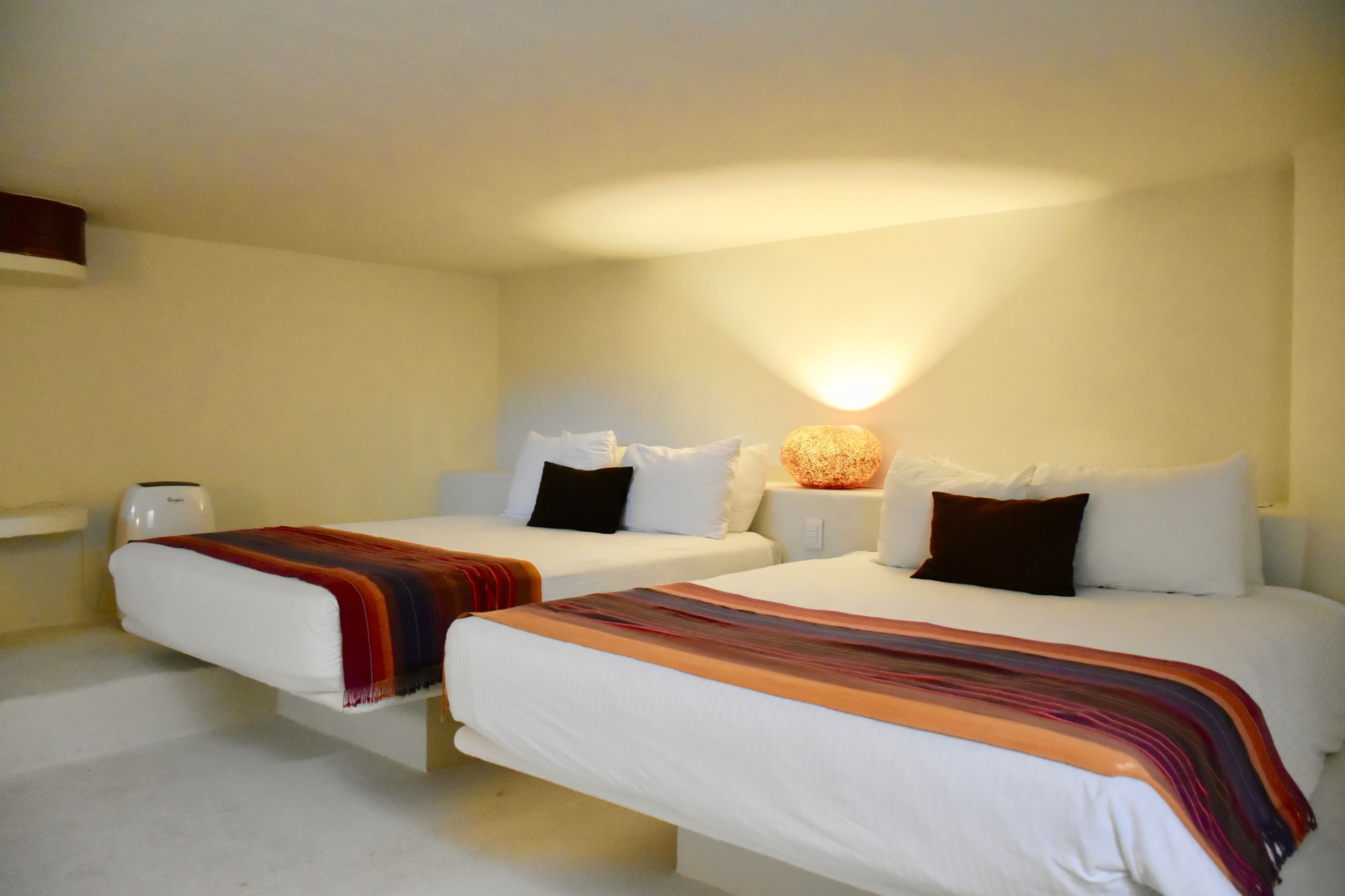 Habitación de hotel con dos camas individuales, cada una con ropa blanca y una manta colorida. Hay una mesita de noche con una lámpara y una pequeña mesa con una taza. La habitación tiene un diseño sencillo y limpio, con una pared de color claro y una lámpara en el techo.