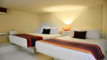 Habitación de hotel con dos camas individuales, cada una con ropa blanca y una manta colorida. Hay una mesita de noche con una lámpara y una pequeña mesa con una taza. La habitación tiene un diseño sencillo y limpio, con una pared de color claro y una lámpara en el techo.