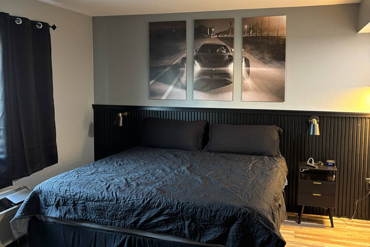 A bedroom with a large bed, a nightstand, a wall-mounted lamp, and a window with curtains.
