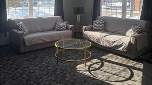 Un salon avec deux canapés, une table basse et une lampe. De grandes fenêtres, ornées de rideaux, se trouvent sur les murs, et le sol est recouvert d’une moquette.