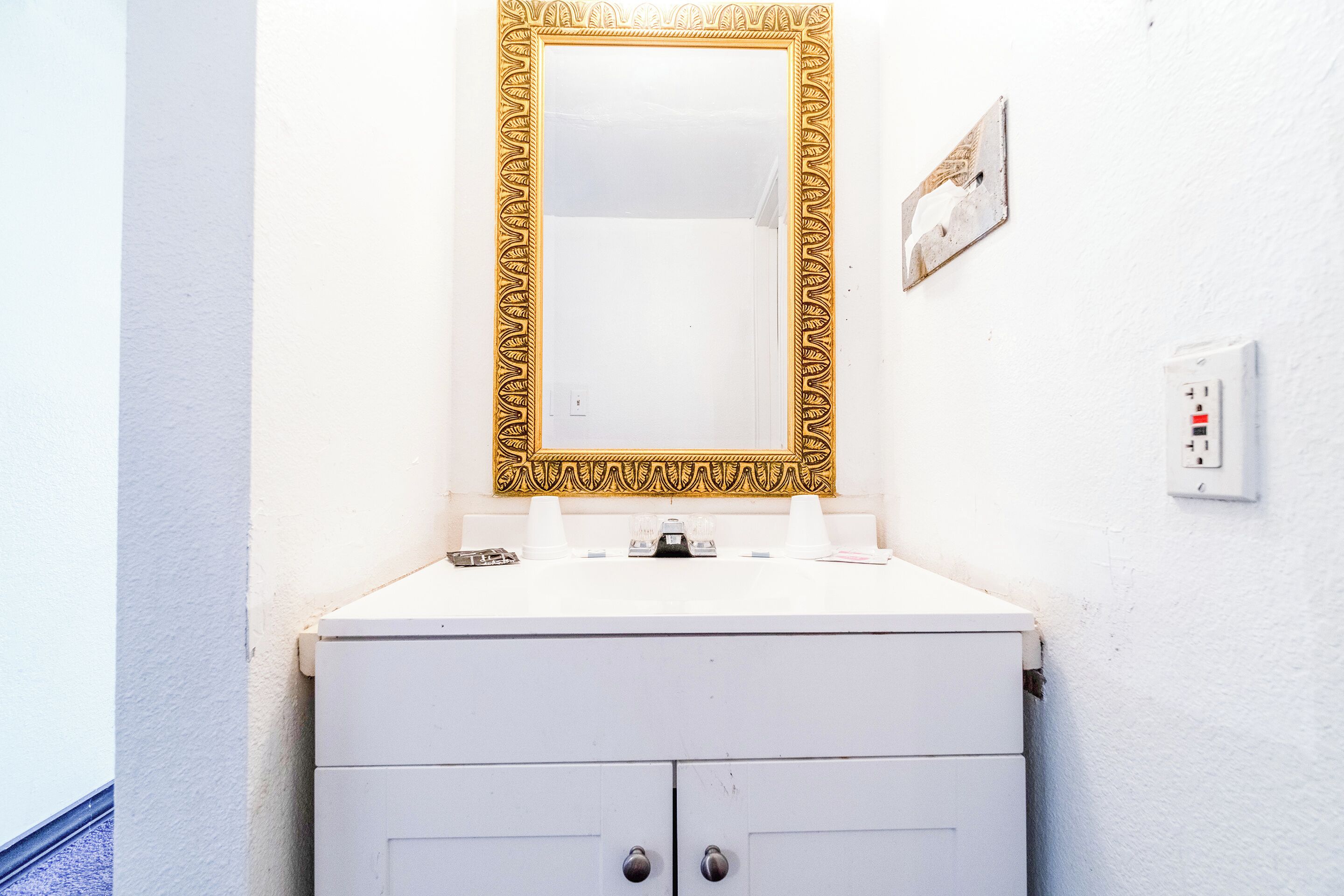 Un baño con lavamanos blanco, un espejo con marco dorado y un enchufe en la pared.