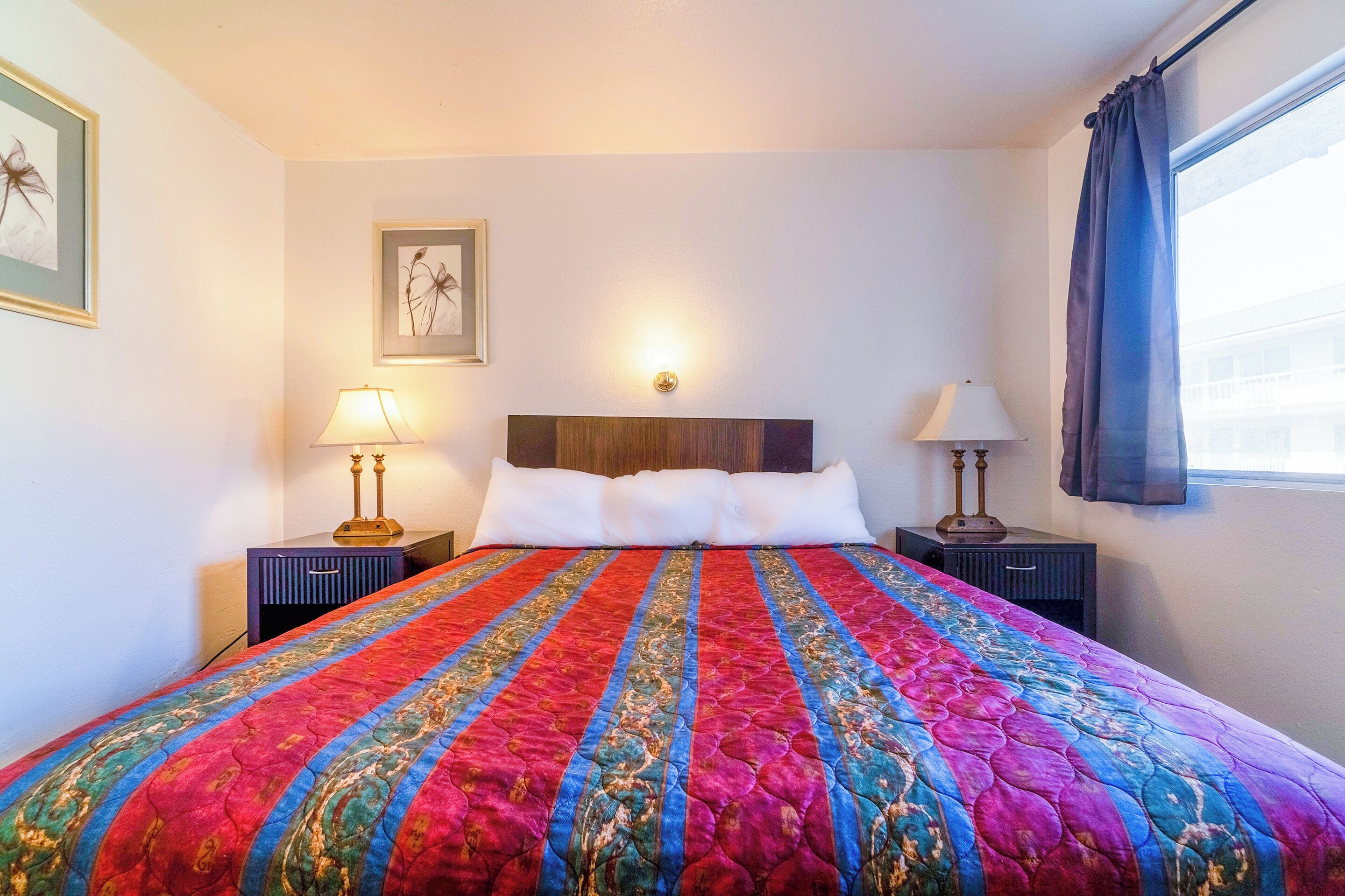 Una cama bien hecha con un colcha colorida, dos lámparas de noche, un cuadro en la pared y una ventana con cortinas.