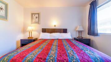 Una cama bien hecha con un colcha colorida, dos lámparas de noche, un cuadro en la pared y una ventana con cortinas.