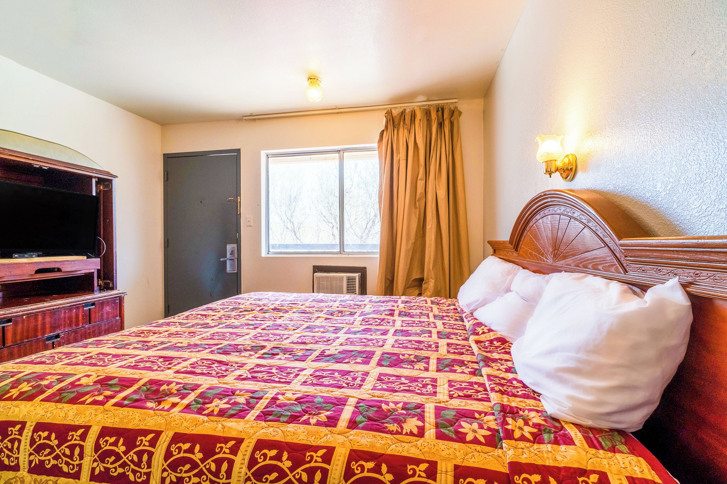 A hotel room with a large bed, a television, a window with curtains, and a wall-mounted light fixture.