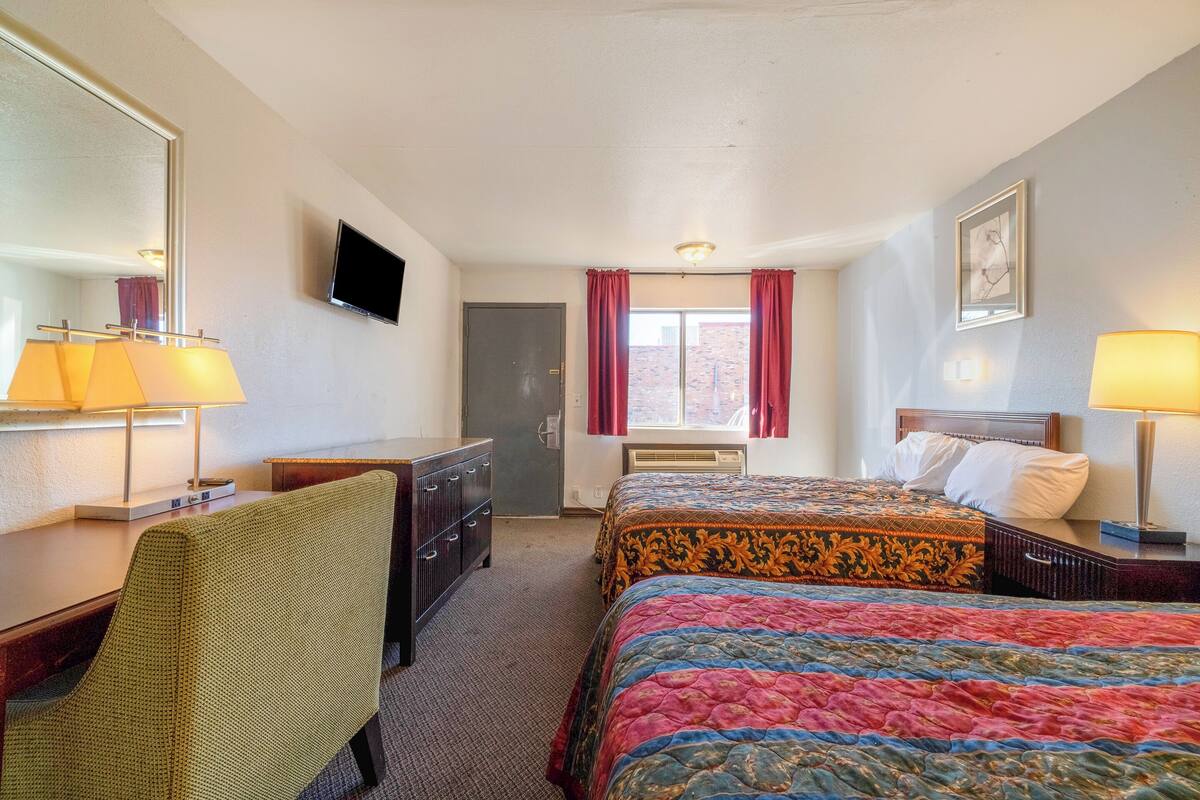 A hotel room with two beds, a desk with a lamp, a chair, a dresser, a TV, and a window with curtains.