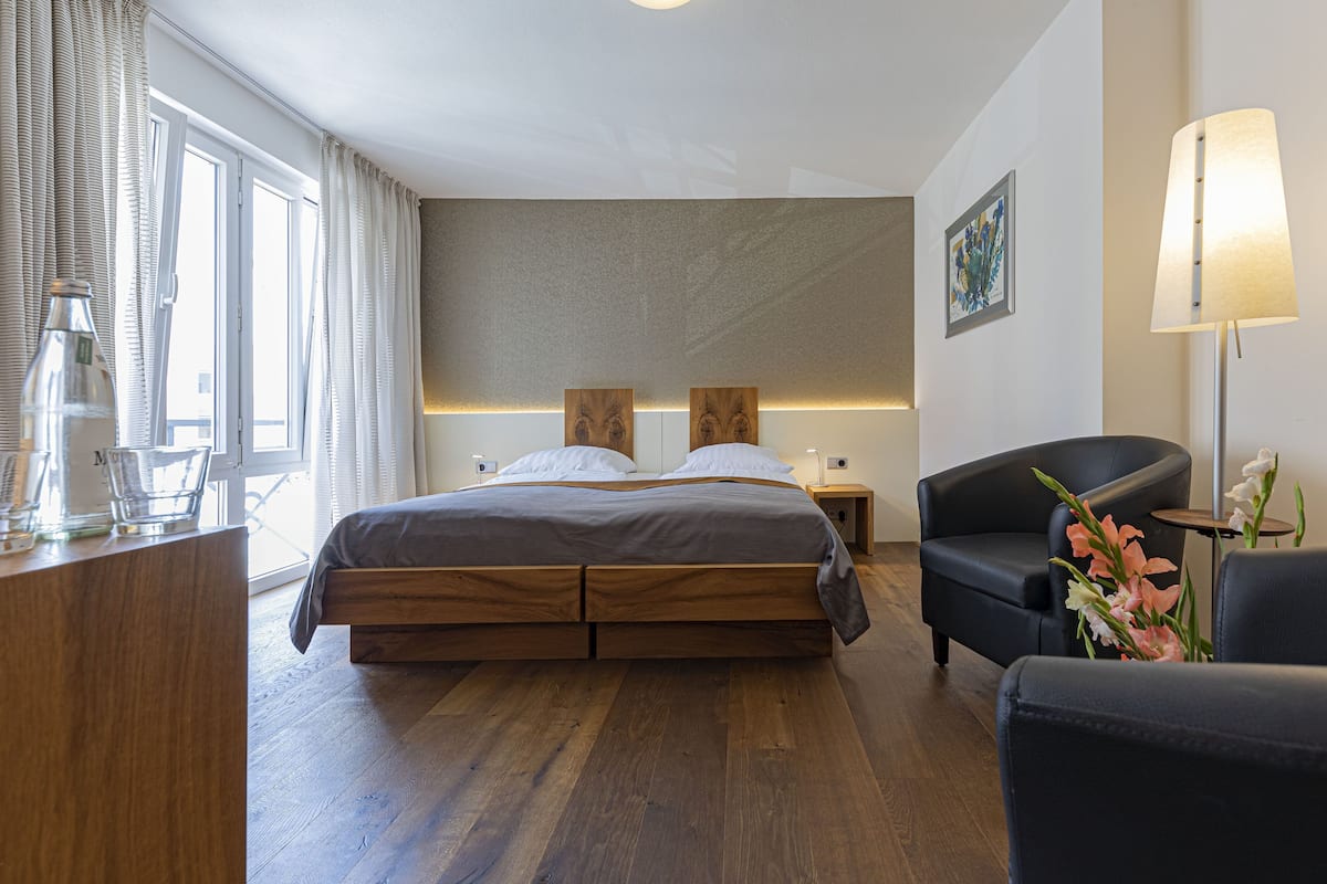 Una habitación de hotel moderna con una cama de madera, un sillón negro, un escritorio con una lámpara y un jarrón de flores.
