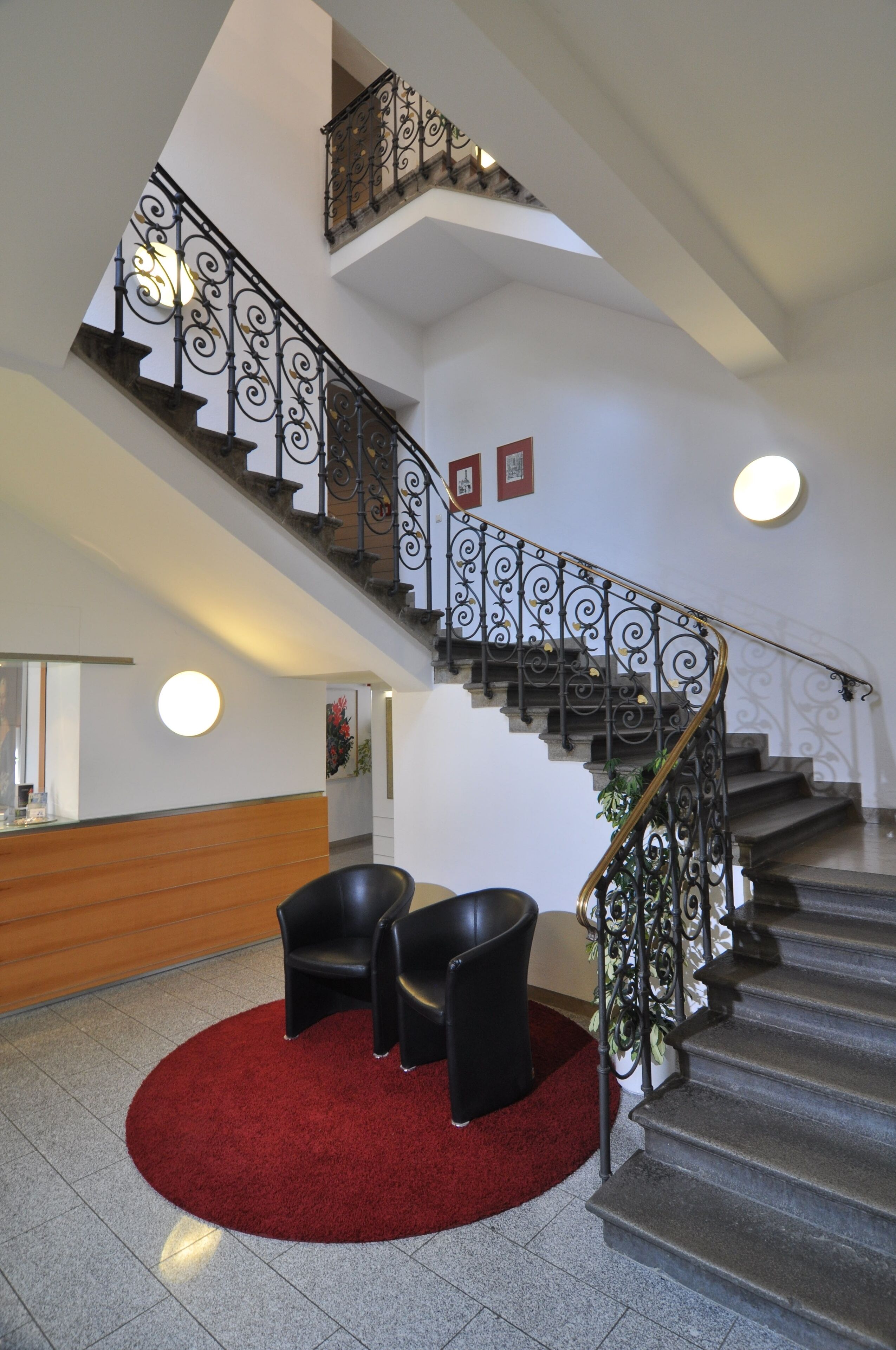 A staircase with ornate metal railings and a red carpet leading to a seating area with two chairs.