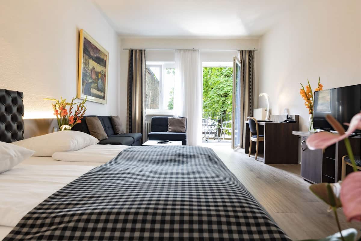 Habitación de hotel con una cama grande, un escritorio, una silla, un televisor y un balcón con vistas a árboles.