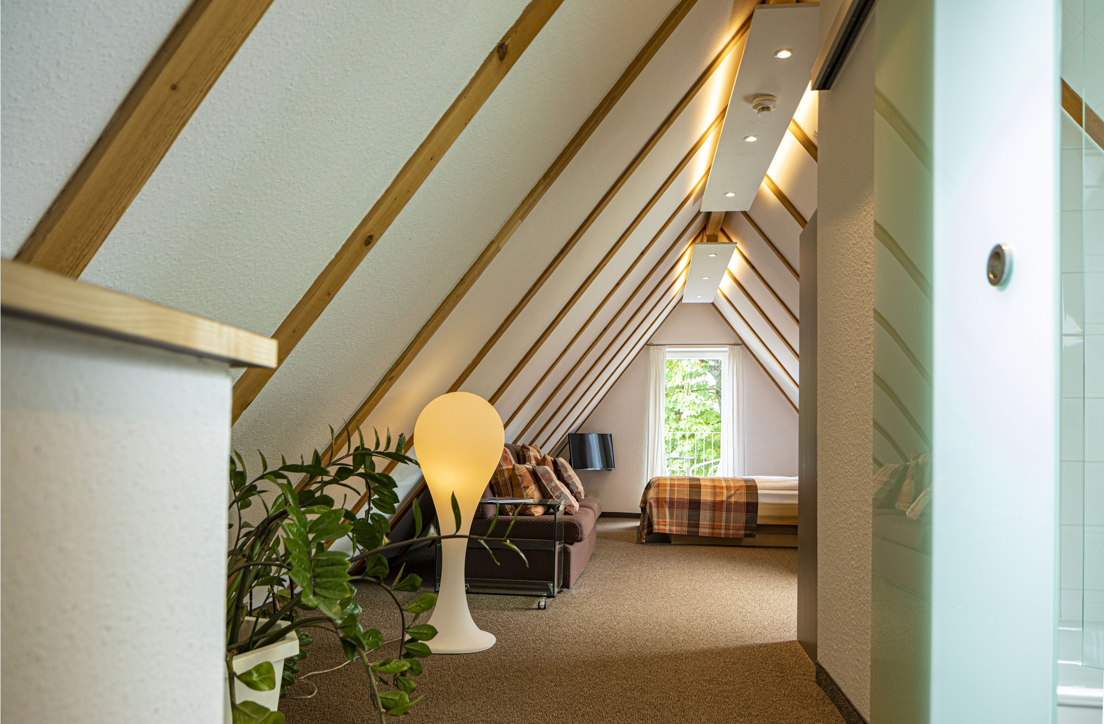 A narrow, sloped ceiling room with a sofa, a lamp, and a bed.