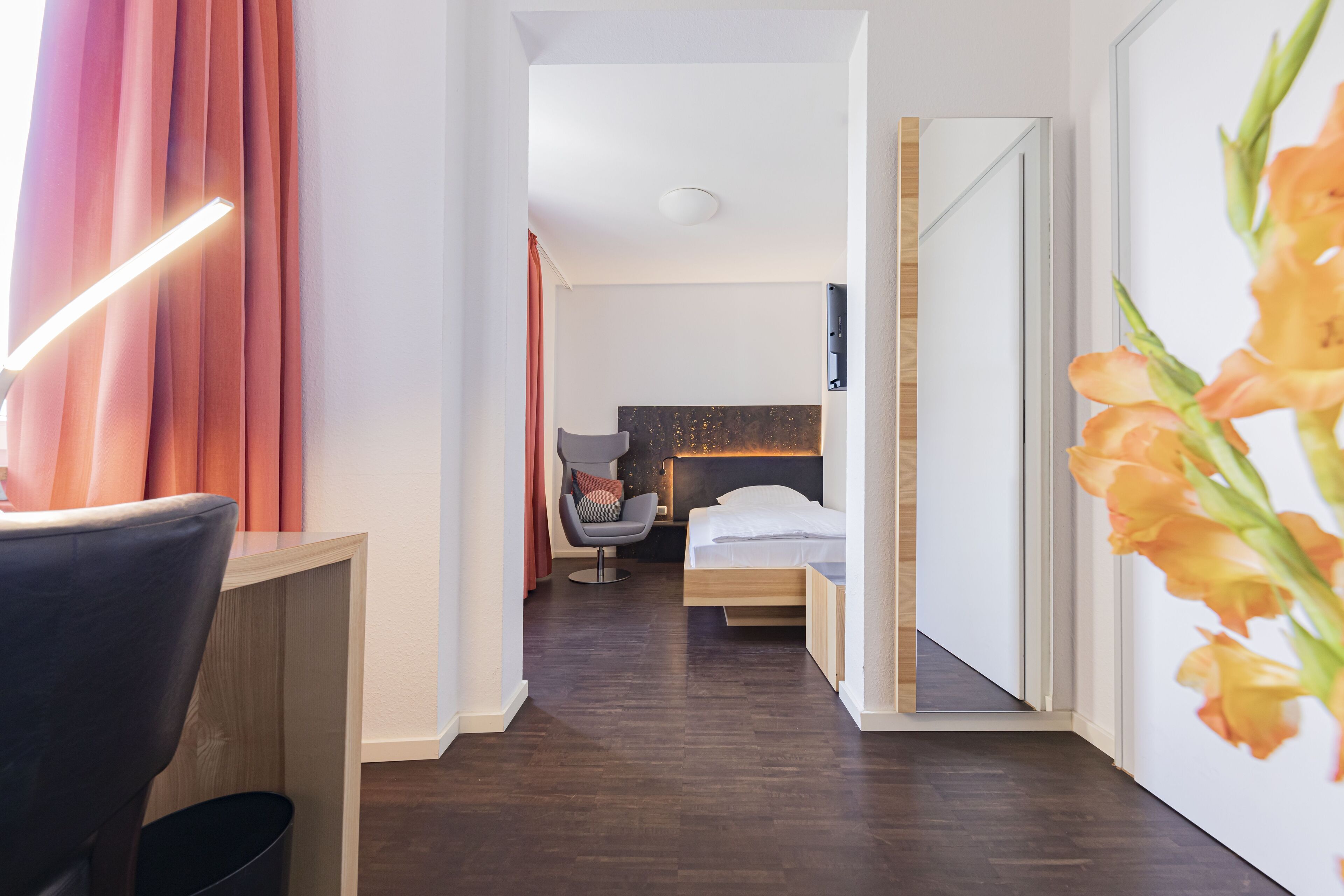 A modern hotel room with a wooden floor, a desk with a chair, a bed with a headboard, and a large mirror.