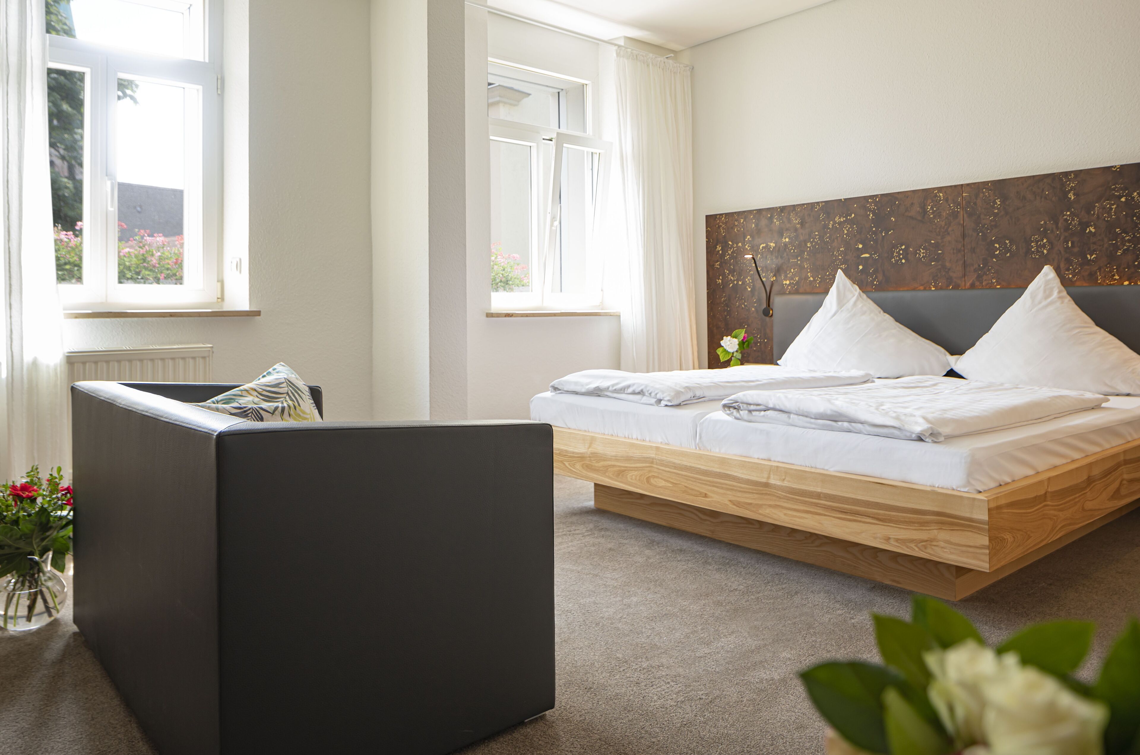 Ein modernes Schlafzimmer mit einem Holzbett, einem dunklen Sofa und Blick auf Blumen durch das Fenster.