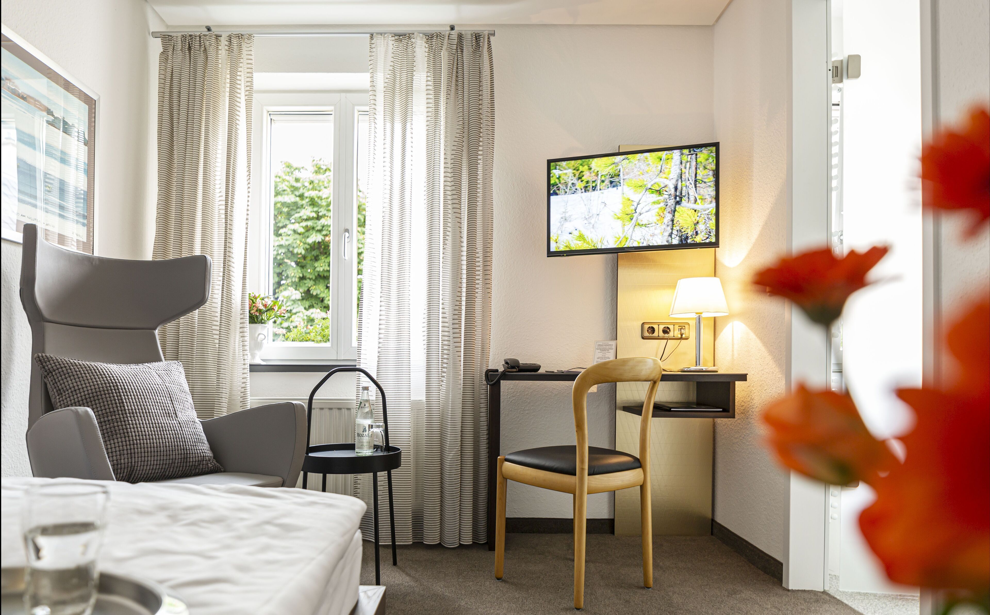 Habitación de hotel con una cama, un escritorio con silla, un televisor montado en la pared y una ventana con cortinas.