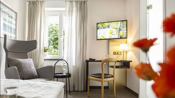 Habitación de hotel con una cama, un escritorio con silla, un televisor montado en la pared y una ventana con cortinas.