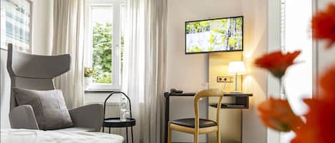 A hotel room with a bed, a desk with a chair, a TV mounted on the wall, and a window with curtains.