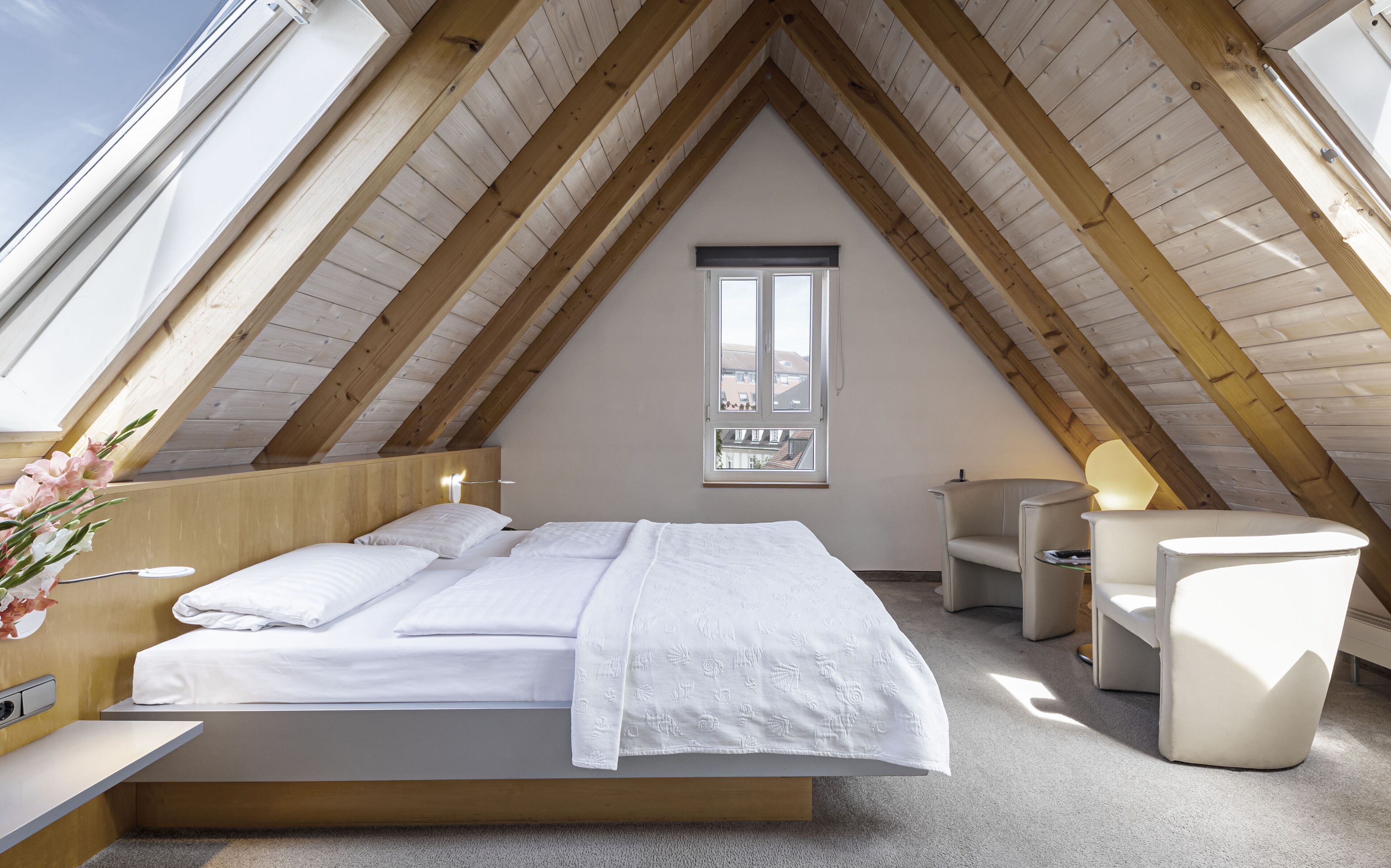 A modern attic bedroom with a sloped wooden ceiling, a large bed with white bedding, a small desk, and a chair.