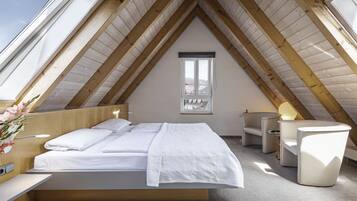 A modern attic bedroom with a sloped wooden ceiling, a large bed with white bedding, a small desk, and a chair.