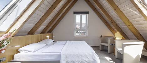 A modern attic bedroom with a sloped wooden ceiling, a large bed with white bedding, a small desk, and a chair.