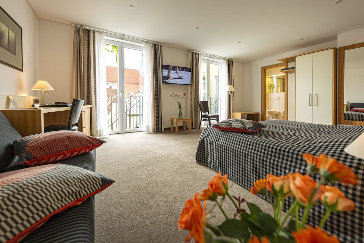 Habitación de hotel con cama, escritorio, silla, televisor y balcón con cortinas.