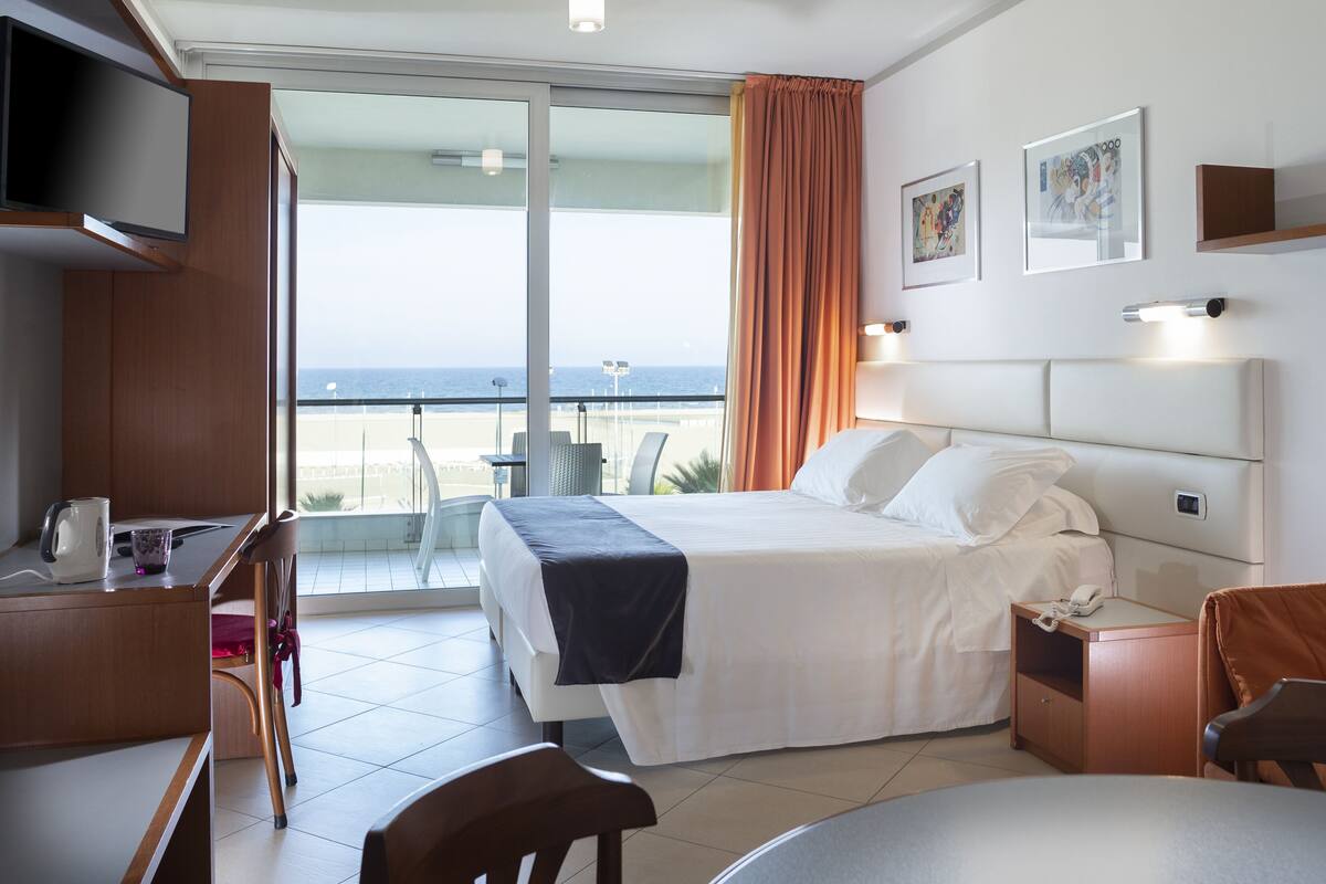 A hotel room with a bed, a desk, a chair, a table, a TV, a balcony with a view of the beach, and artwork on the wall.