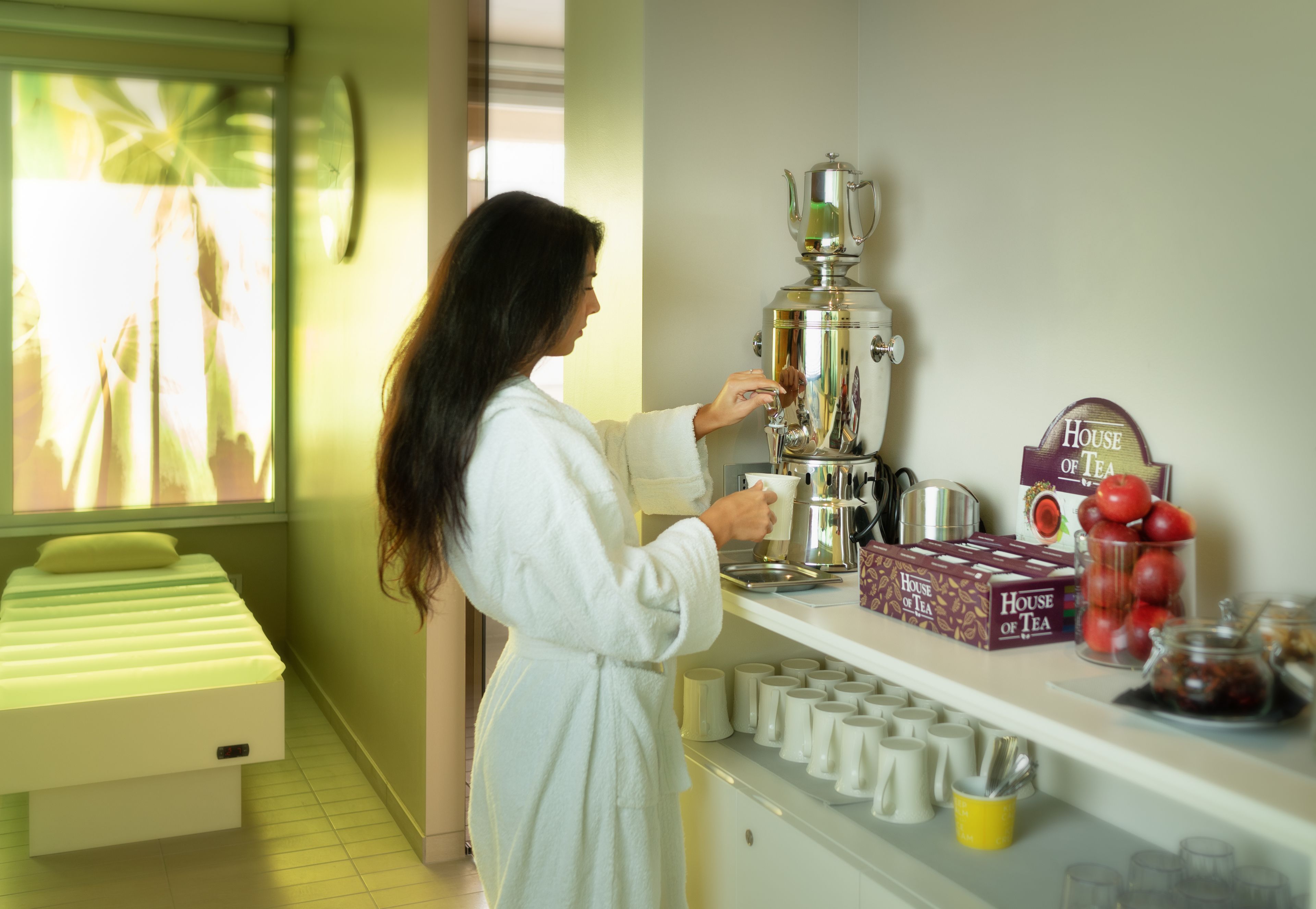 A person in a white robe preparing tea with a coffee pot in a room with a bed, tea boxes, and a window with a view.