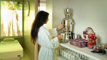 A person in a white robe preparing tea with a coffee pot in a room with a bed, tea boxes, and a window with a view.