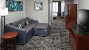 Una habitación de hotel moderna con un sofá seccional gris, una mesita de madera, un televisor de pantalla plana y una alfombra estampada.