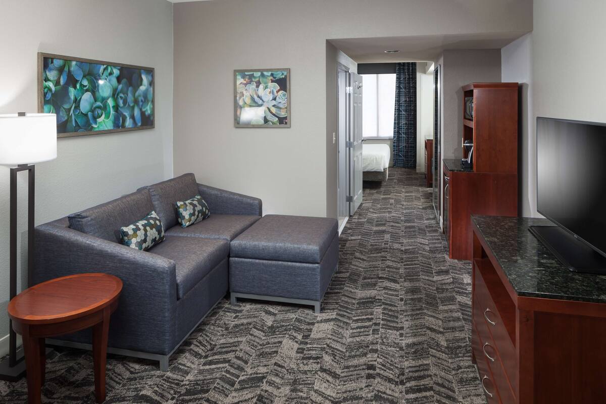 A modern hotel room with a grey sectional sofa, a small wooden side table, a flat-screen TV, and a patterned carpet.