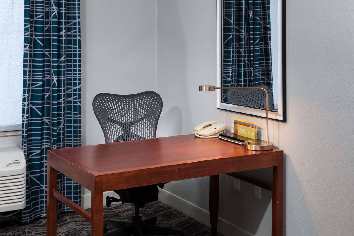 A hotel room with a wooden desk, a chair, a telephone, and a framed picture on the wall.