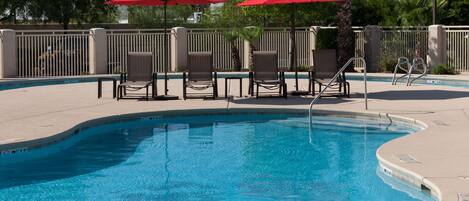 A swimming pool with lounge chairs and red umbrellas, surrounded by a fence and trees.