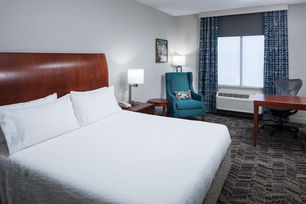 A hotel room with a large bed, a desk, a chair, a lamp, and an air conditioning unit.