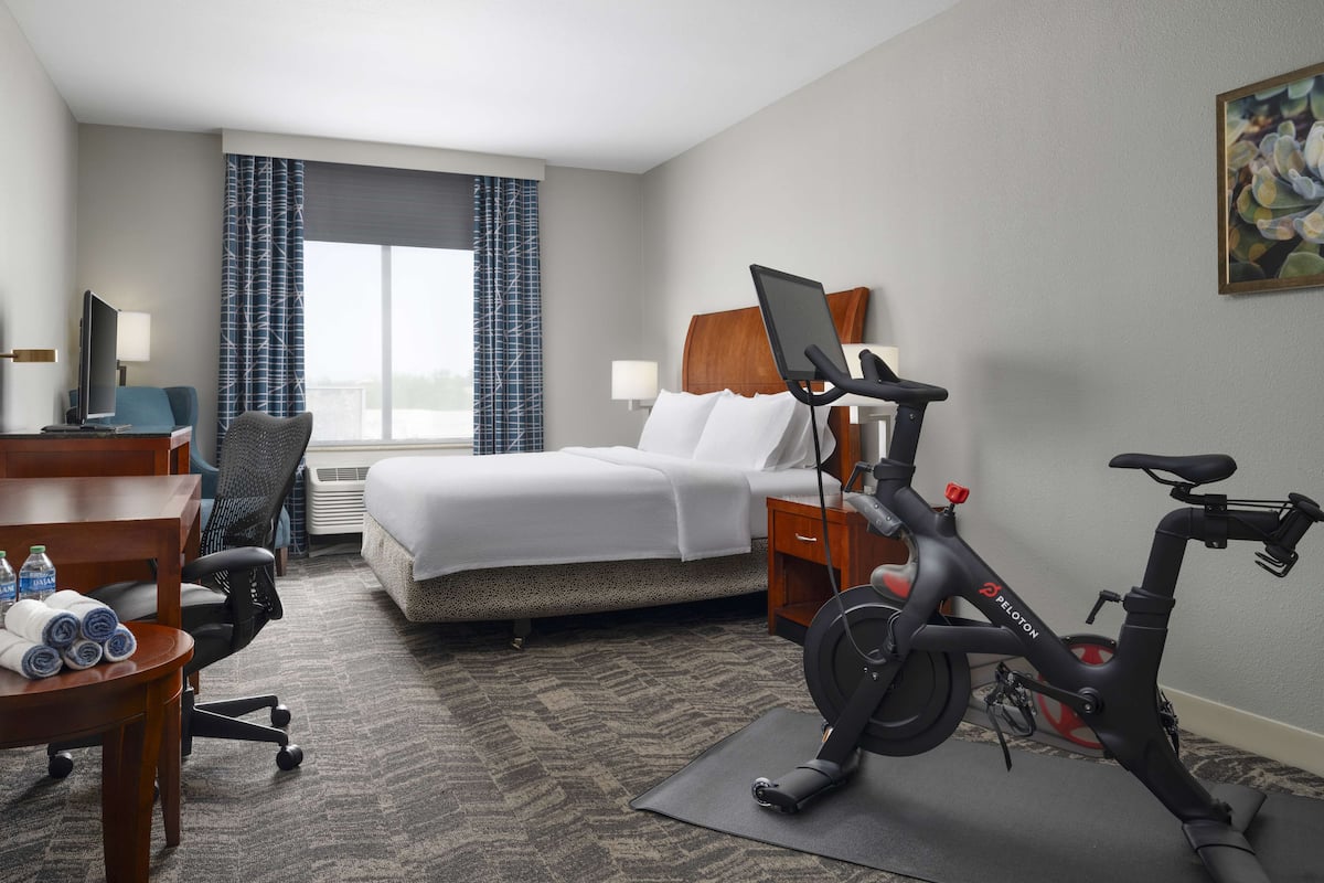 A hotel room with a bed, a desk with a computer, a chair, a treadmill, and a window with curtains.