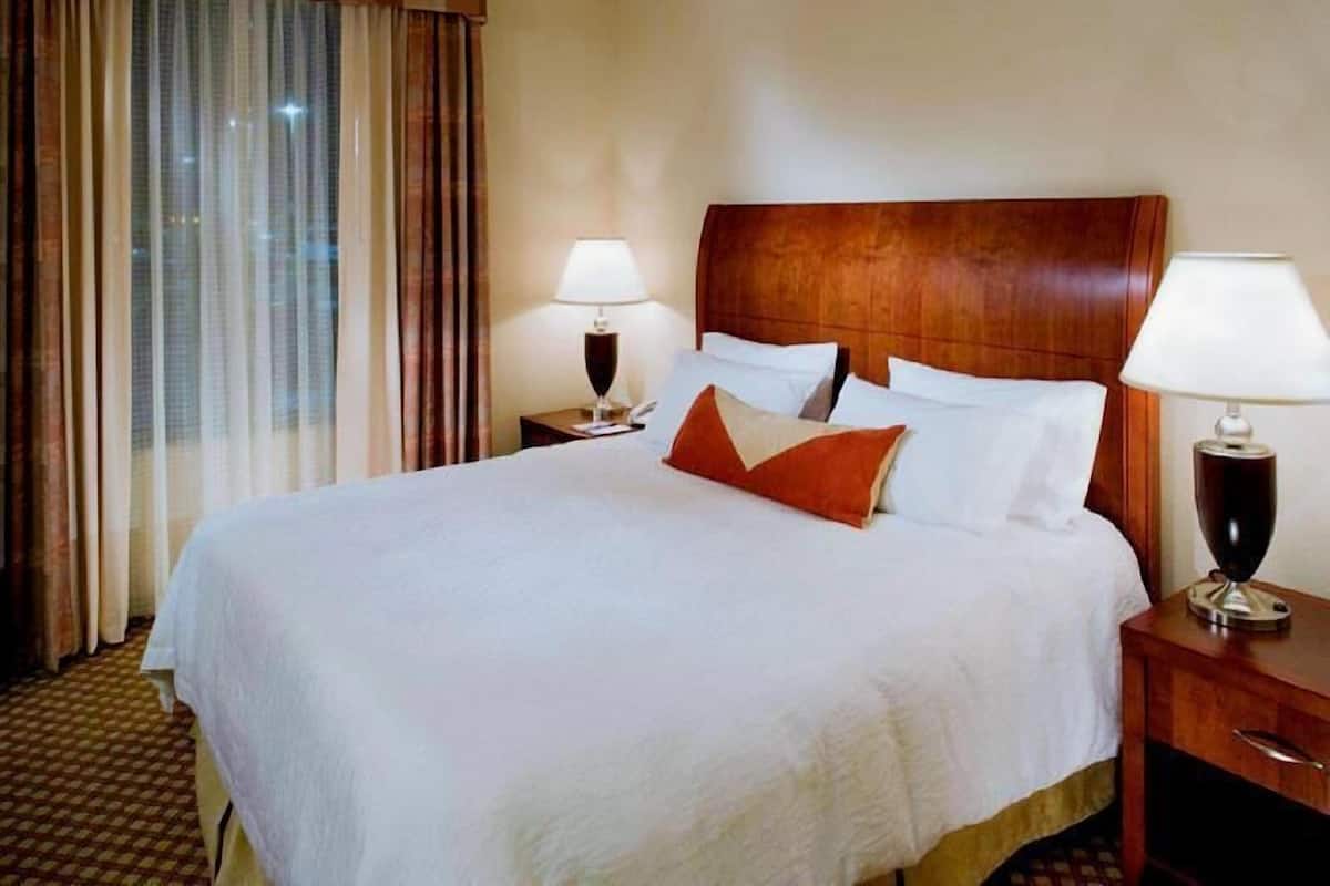 A hotel room with a large bed, two bedside tables with lamps, a wooden headboard, and a view of the city through curtains.