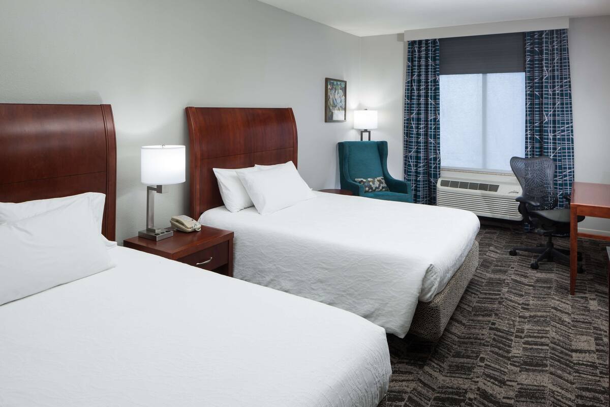 A hotel room with two beds, a desk, a chair, a lamp, and a window with curtains.